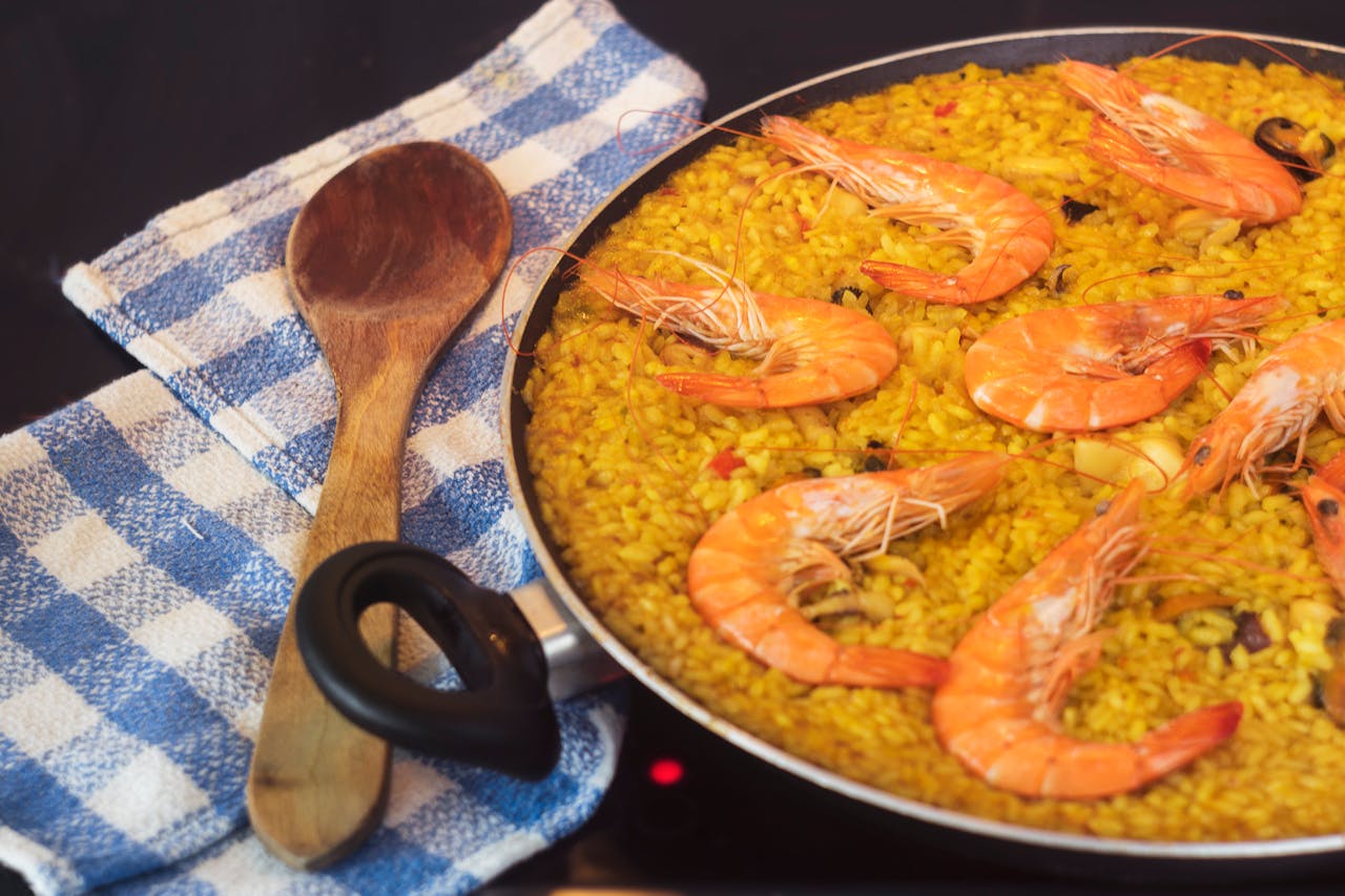 heros-img Close-up of a seafood paella with prawns in a pan, a wooden spoon, and blue towel.